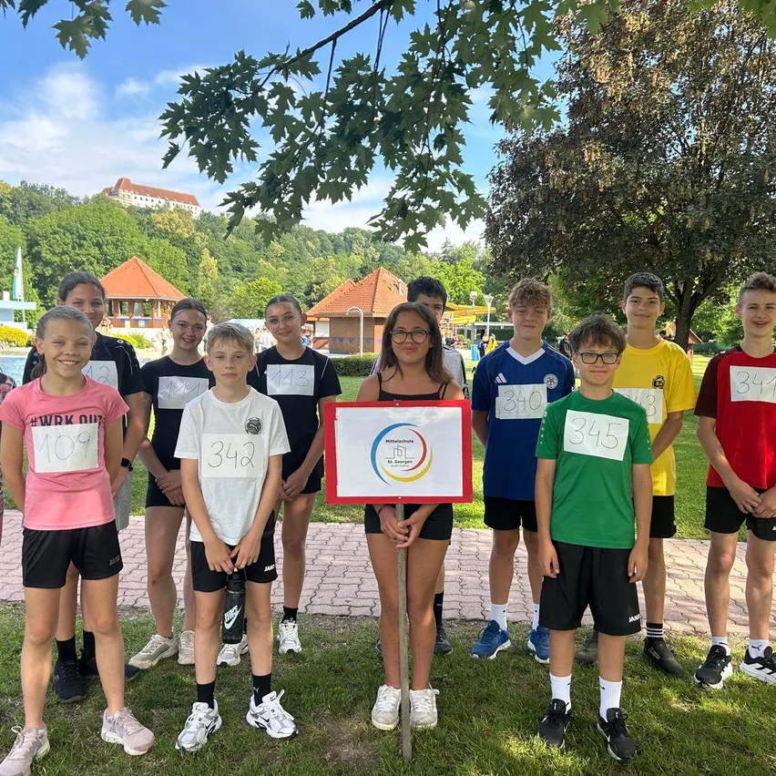 Eine Gruppe junger Athleten posiert auf einem Rasenfeld mit Nummern auf ihren Trikots für ein Foto. Ein Mädchen hält ein Schild mit Logo und Nummer 342. Hinter ihnen stehen Gebäude und Bäume.