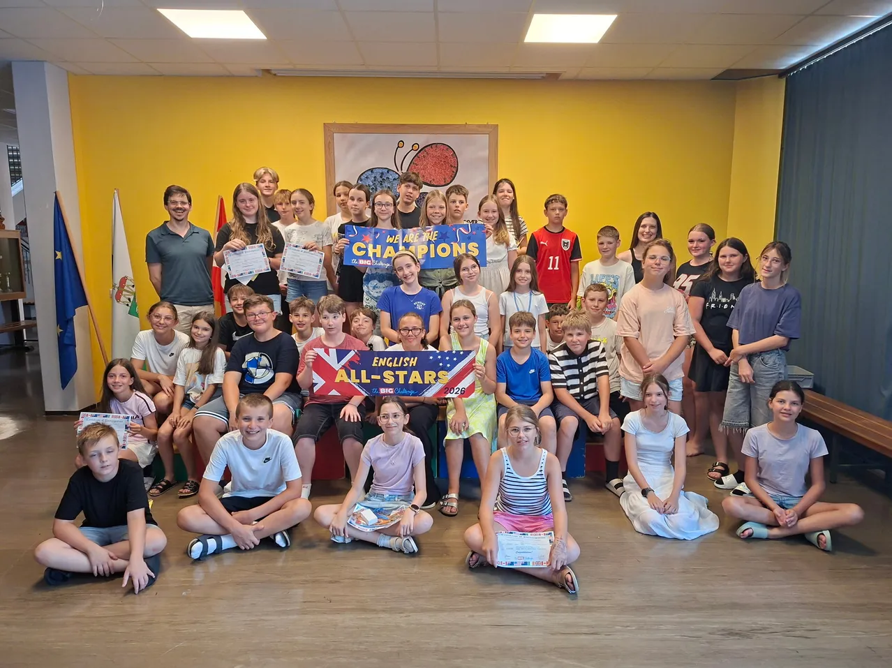 Eine Gruppe von Kindern und Erwachsenen posiert für ein Foto mit einem Banner mit der Aufschrift 'We are the Champions'. Sie befinden sich in einem Raum mit gelben Wänden.