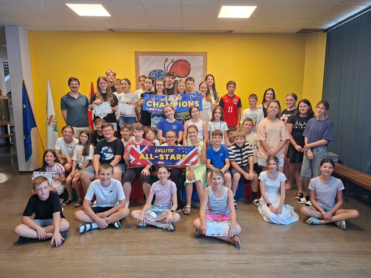 A group of children and adults posing for a photo with a banner reading 'We are the Champions'. They are in a room with yellow walls.