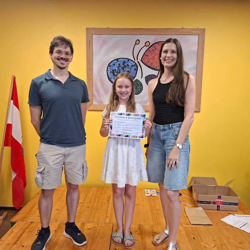 A young girl stands on a stage holding a certificate of participation. Two adults stand beside her, smiling, with a Canadian flag and a butterfly painting in the background.