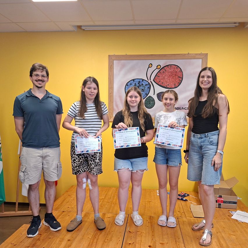 Five people are standing on a stage, holding certificates. One person is a man and the others are women. They are smiling for the camera. The room is yellow.