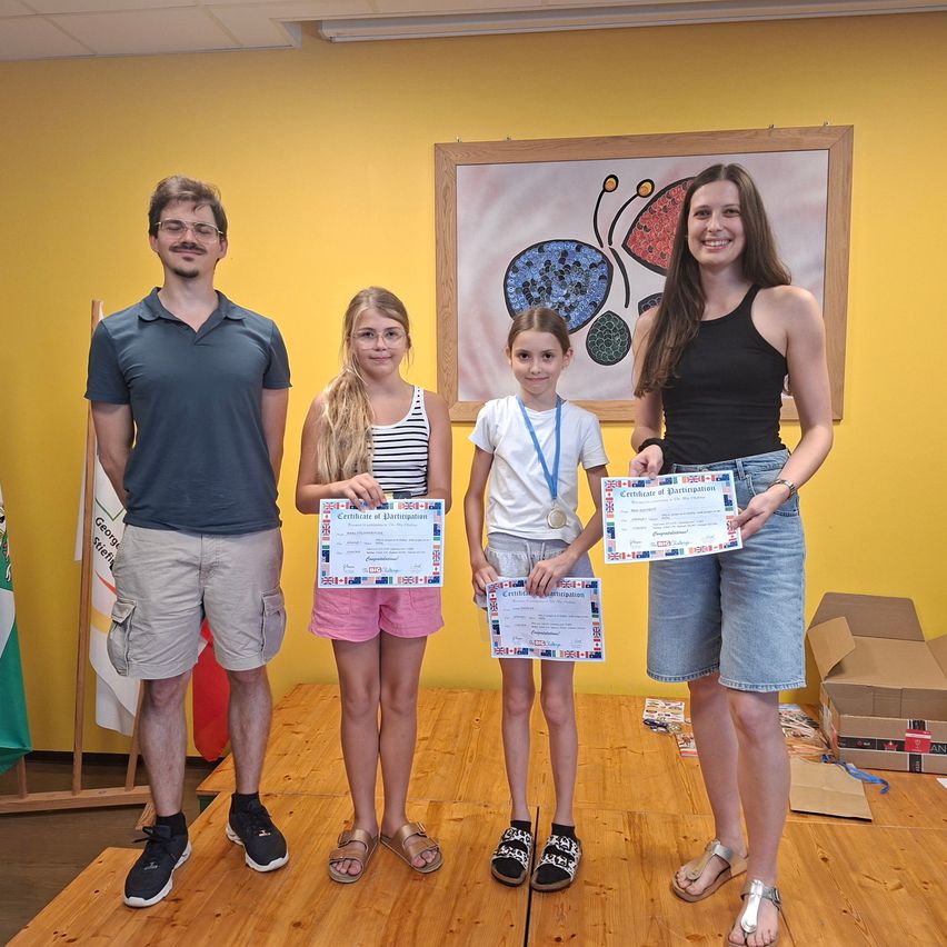 A man and three girls are on a stage holding certificates. The girls wear glasses, sandals, and medals. A yellow wall with a painting and flags is behind them.