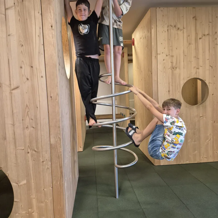 Drei Jungen klettern auf einer spiralförmigen Metallstruktur in einem hölzernen Spielzimmer. Ein Junge hängt an der Spirale, während die anderen beiden hochklettern.