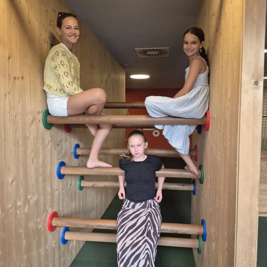 Drei junge Mädchen klettern auf einer Holzleiter in einem Spielbereich. Das Mädchen in der Mitte trägt ein schwarzes Shirt und Zebra-Print-Hose.