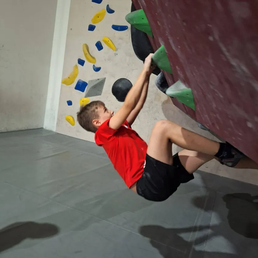 Ein junger Junge in einem roten T-Shirt und schwarzen Shorts klettert an einer Wand mit verschiedenen farbigen Griffen. Er hängt kopfüber und hält die Wand mit Händen und Füßen fest.