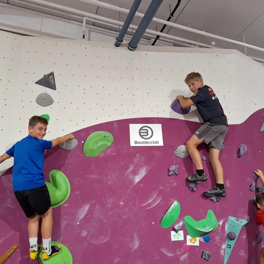 Zwei Jungen klettern an einer Wand mit verschiedenen farbigen Griffen. Ein Junge trägt ein blaues Shirt, der andere ein schwarzes. Die Wand ist in Rosa und Weiß gestrichen, und es gibt ein Logo an der Wand.