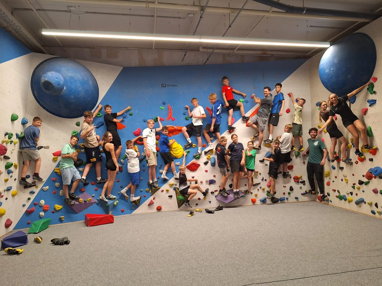 Eine Gruppe junger Kletterer posiert für ein Foto in einem Kletterzentrum, mit bunten Klettergriffen an der Wand und zwei großen blauen Kugeln, die von der Decke hängen.