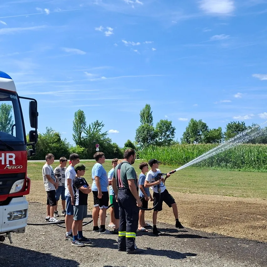 Eine Gruppe junger Jungen beobachtet, wie ein Feuerwehrmann den Einsatz eines Feuerwehrschlauchs demonstriert. Sie stehen auf einem Kiesweg, neben einem geparkten Feuerwehrwagen.