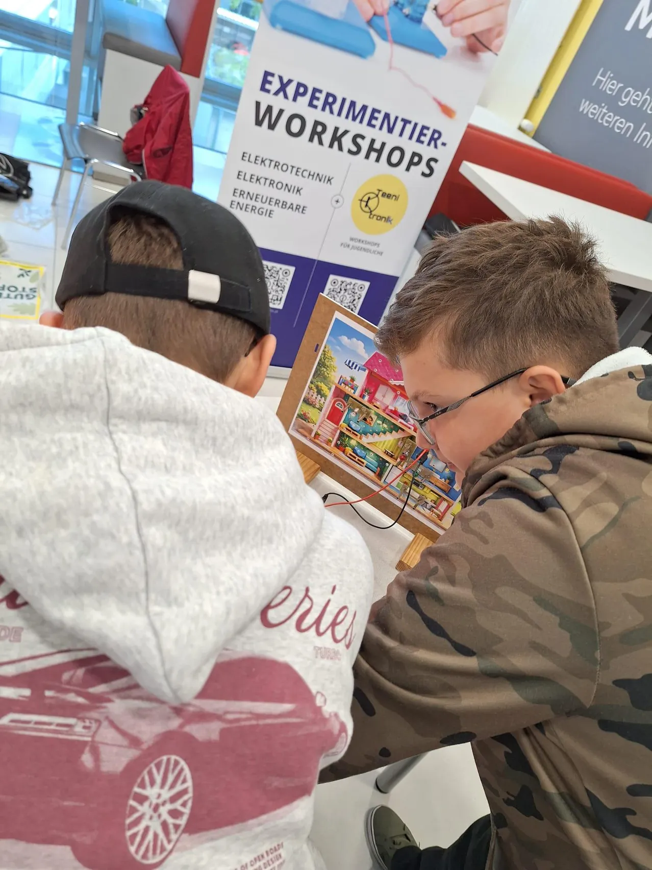 Zwei junge Jungen arbeiten in einem Raum an einem Projekt. Ein Junge trägt eine graue Kapuzenjacke mit einem roten Autodesign und einer schwarzen Kappe, während der andere eine Tarnjacke und eine Brille trägt. Sie schauen beide auf ein kleines Brett mit einem Hausdesign.