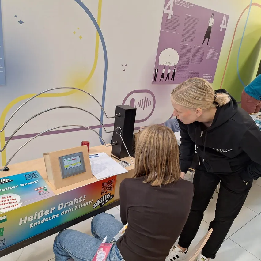 Eine Frau sitzt an einem Schreibtisch mit einem Monitor und einem Lautsprecher, während eine andere Frau steht und den Monitor betrachtet. Es gibt ein Poster mit dem Text 'HeiBer Draht! Entdecke dein Talent' auf dem Schreibtisch.