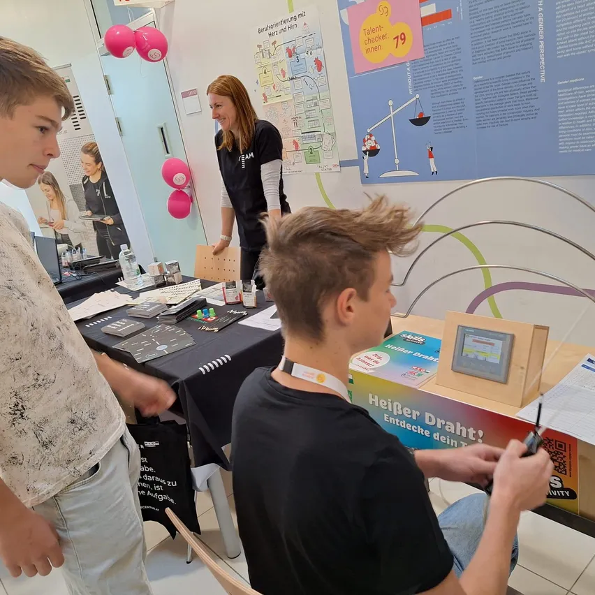 Zwei Jungen sind in einem Labor an einem wissenschaftlichen Experiment beteiligt. Ein Junge sitzt und arbeitet an einem Gerät, während der andere Junge steht und das Experiment beobachtet. Eine Frau steht hinter ihnen und beobachtet das Experiment lächelnd.