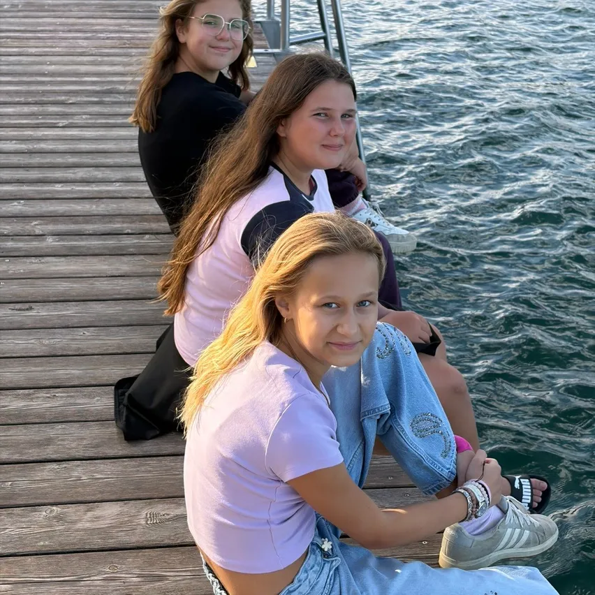 Drei junge Mädchen sitzen auf einem Holzsteg am Wasser. Das Mädchen in der Mitte trägt eine Brille und eine Halskette. Das Mädchen auf der rechten Seite trägt ein Armband und Turnschuhe.