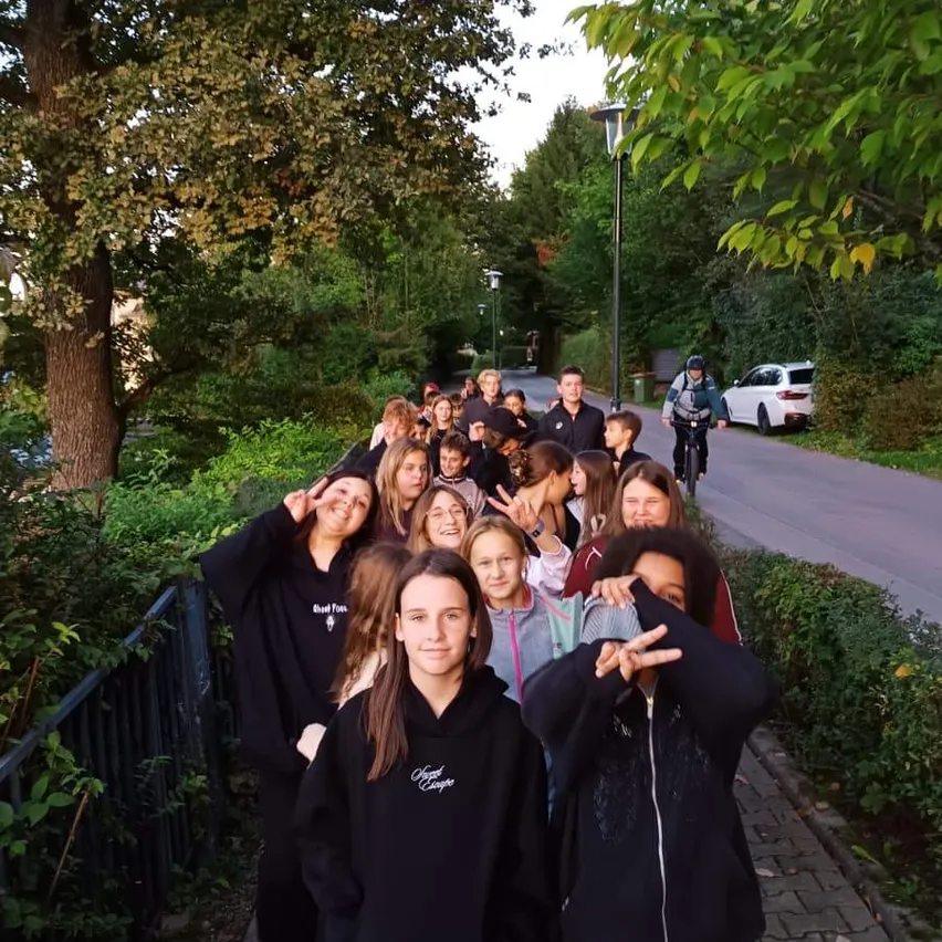 Eine Gruppe von Menschen, hauptsächlich junge Erwachsene, steht in einer Reihe auf einem gepflasterten Gehweg und posiert für ein Foto. Sie sind in schwarzen Kapuzenpullovern und Hosen gekleidet, einige tragen Turnschuhe. Der Hintergrund zeigt Bäume, einen Metallzaun und eine Straße mit einem weißen Auto und einer Person, die mit dem Fahrrad fährt.