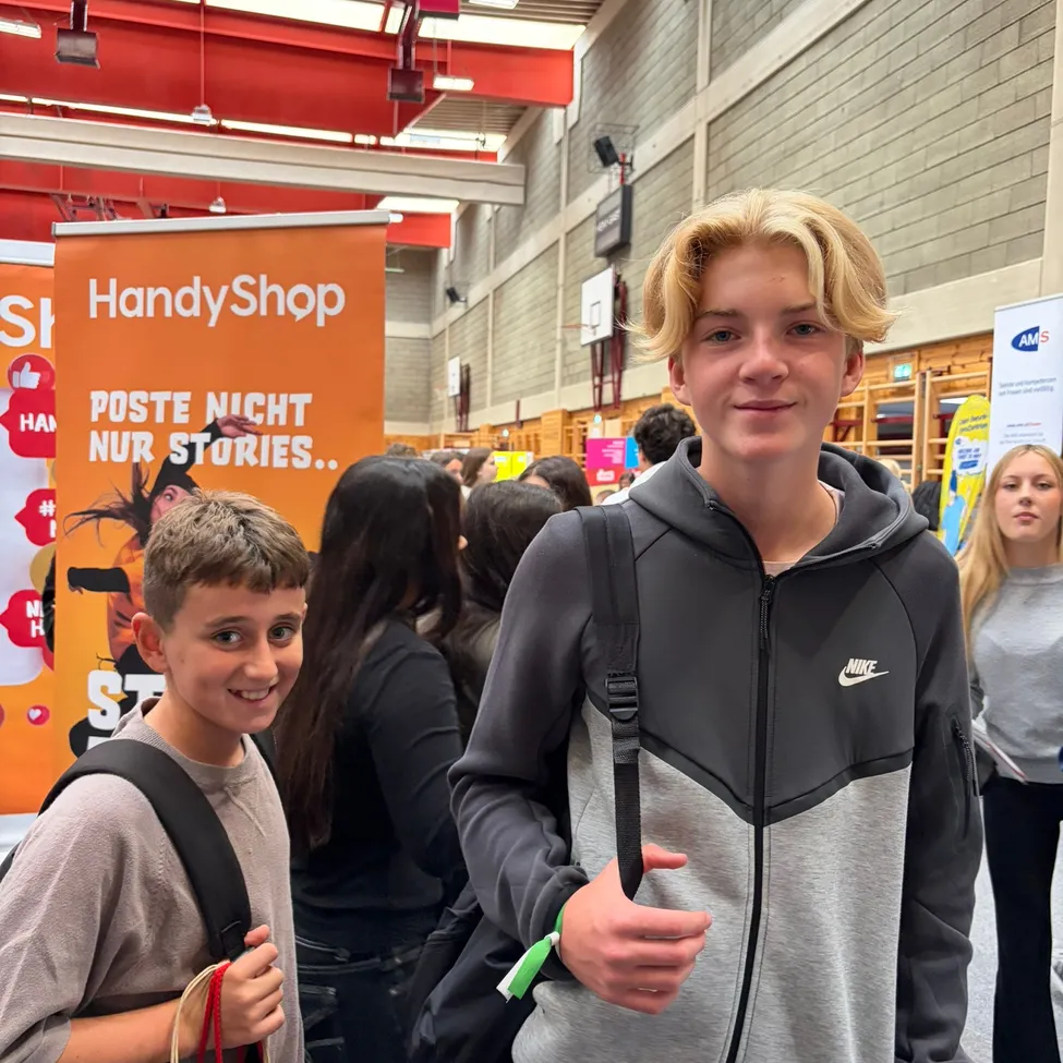 Zwei Jungen stehen in einem Flur, einer trägt eine graue Kapuzenjacke mit Nike-Logo und der andere trägt einen braunen Papierbeutel. Hinter ihnen steht eine Menschenmenge und ein Banner mit dem Text 'HandyShop' und 'Poste nicht nur stories..'