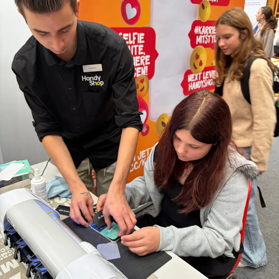 Ein Mann und eine Frau arbeiten an einem Projekt an einem Tisch. Der Mann trägt ein schwarzes Shirt mit dem Wort 'Handy' darauf. Die Frau trägt eine graue Kapuzenjacke.