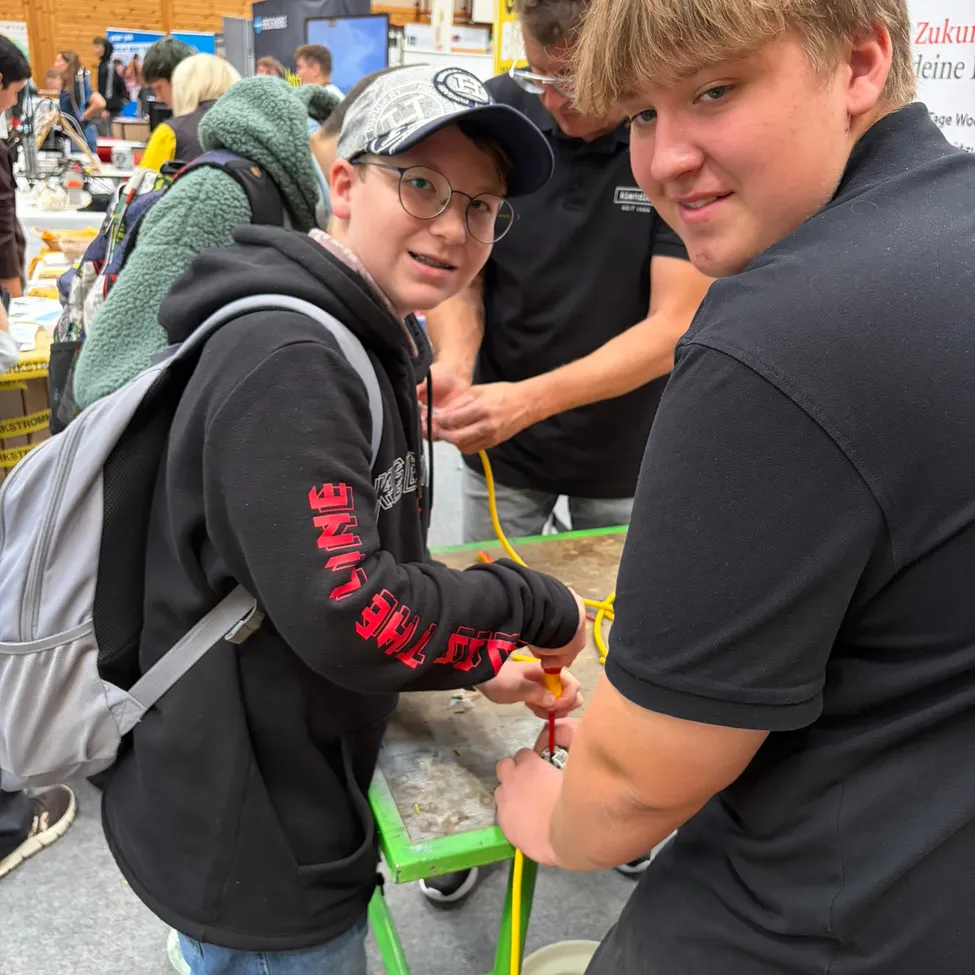 Zwei Jungen arbeiten an einem Projekt an einem Tisch in einem geschäftigen Innenbereich. Der Junge auf der linken Seite trägt eine Brille und einen Rucksack, während der Junge auf der rechten Seite eine Kappe trägt und ein Werkzeug in der Hand hält.