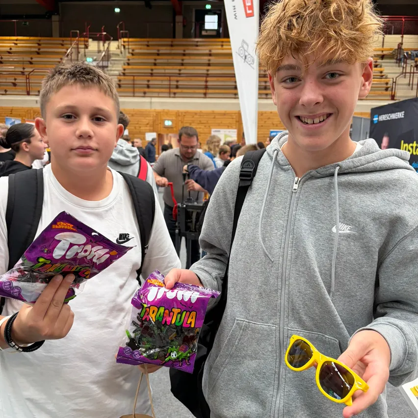 Zwei junge Jungen stehen in einem großen Innenbereich, möglicherweise einem Kongresszentrum. Ein Junge hält eine Tüte Süßigkeiten und der andere eine gelbe Sonnenbrille. Sie lächeln beide und schauen in die Kamera.