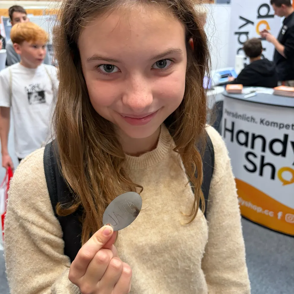 Ein junges Mädchen mit langen braunen Haaren lächelt und hält ein kleines Metallobjekt in der Hand. Sie trägt einen beigen Pullover und einen schwarzen Rucksack. Im Hintergrund sind verschwommene Menschen und ein Schild zu sehen, das 'Handy Shop' besagt.