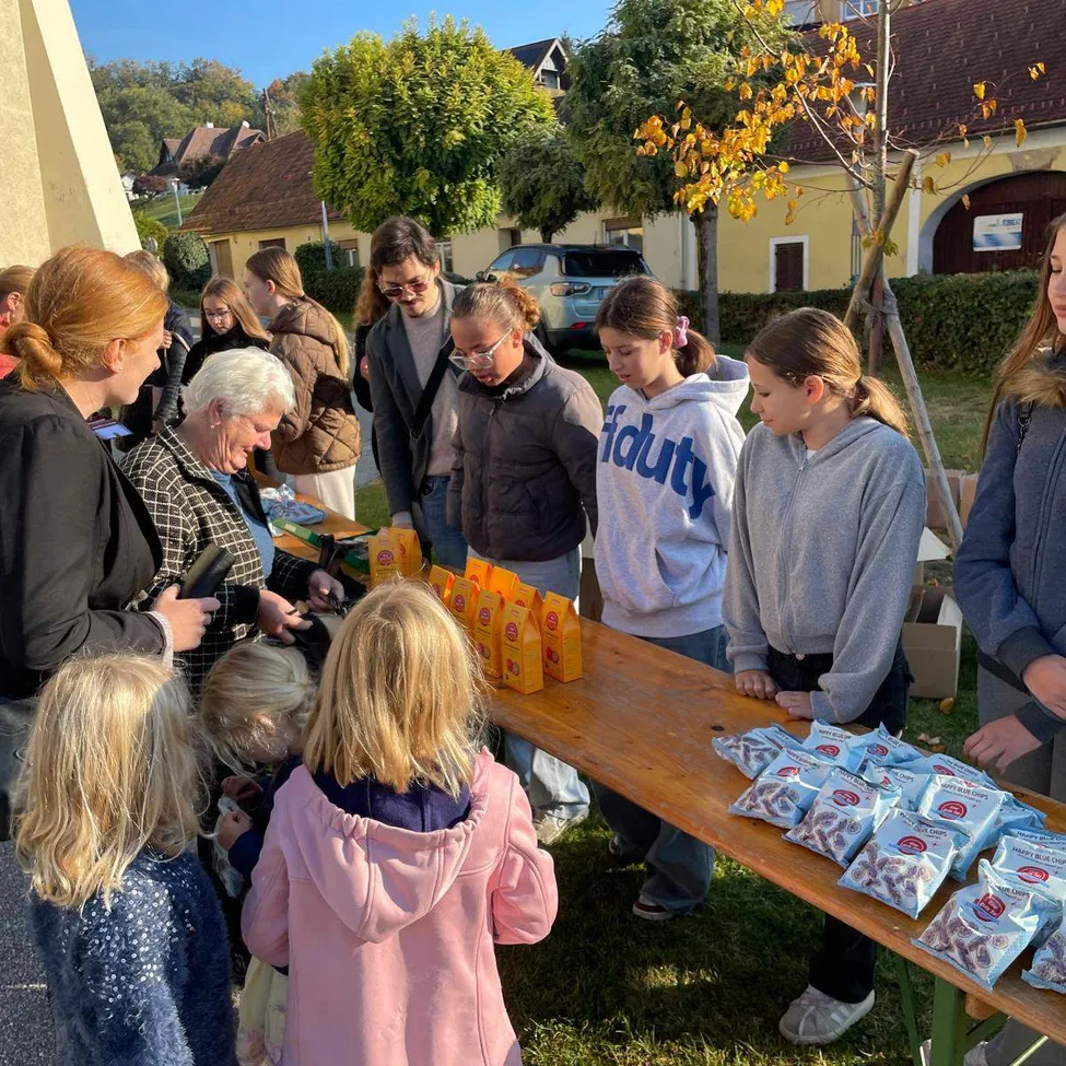 Eine Gruppe von Menschen, einschließlich Erwachsener und Kindern, versammelt sich um einen Tisch mit Essensartikeln. Der Tisch steht im Freien, mit einem Rasenbereich und einigen Bäumen im Hintergrund.