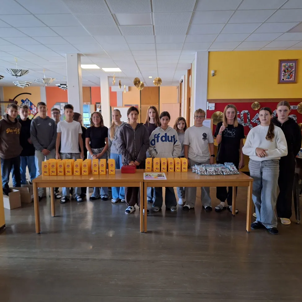 Eine Gruppe junger Leute steht um zwei Tische mit gelben Boxen herum.