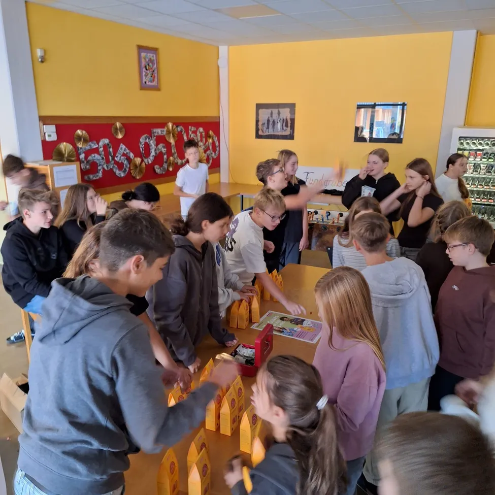 Eine Gruppe von Schülern versammelt sich um einen Tisch in einem Klassenzimmer, einige schauen auf eine Tafel mit Musiknoten und andere schauen in eine Box mit Snacks.
