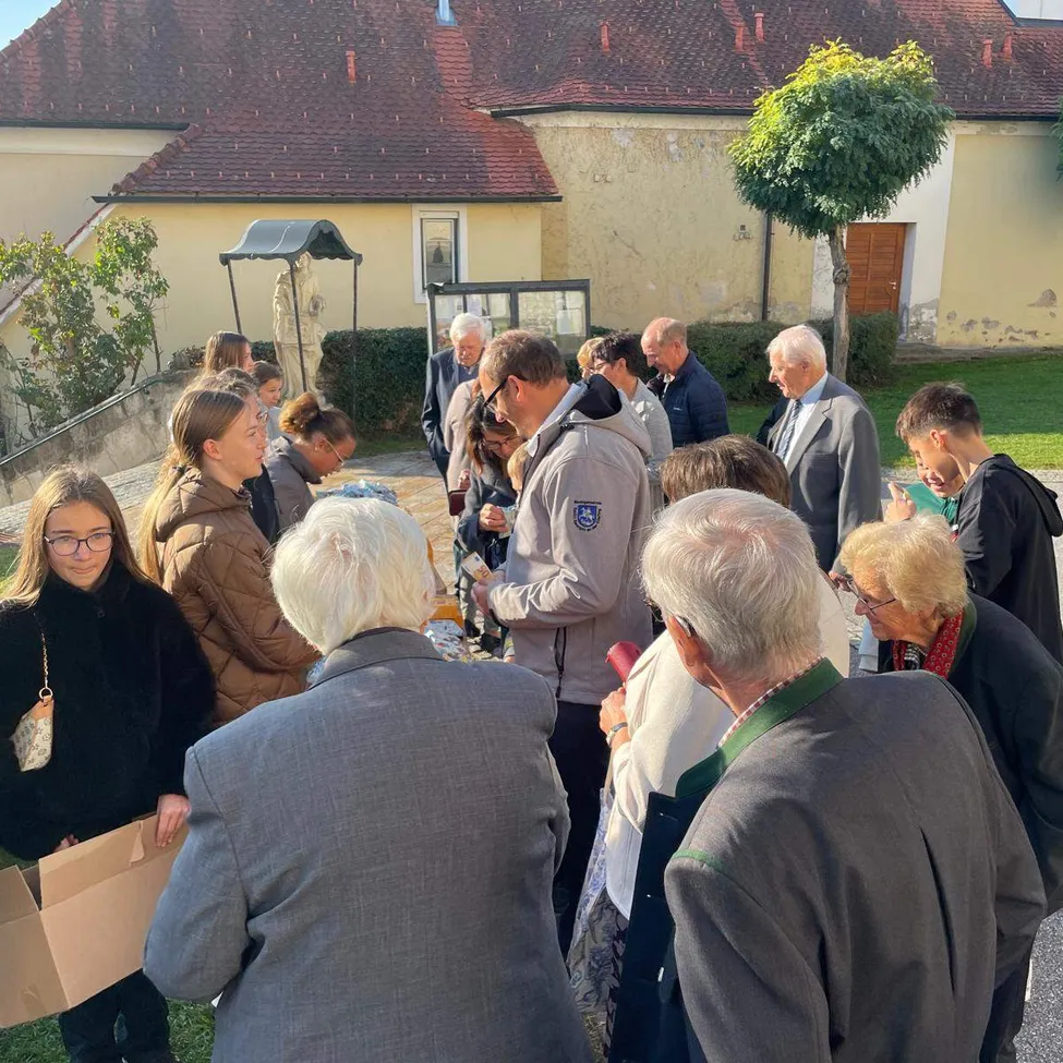 Eine Gruppe von Menschen versammelt sich vor einem Gebäude, wahrscheinlich für eine Gemeinschaftsveranstaltung. Einige tragen Brillen und Jacken, und einige halten Papiere.