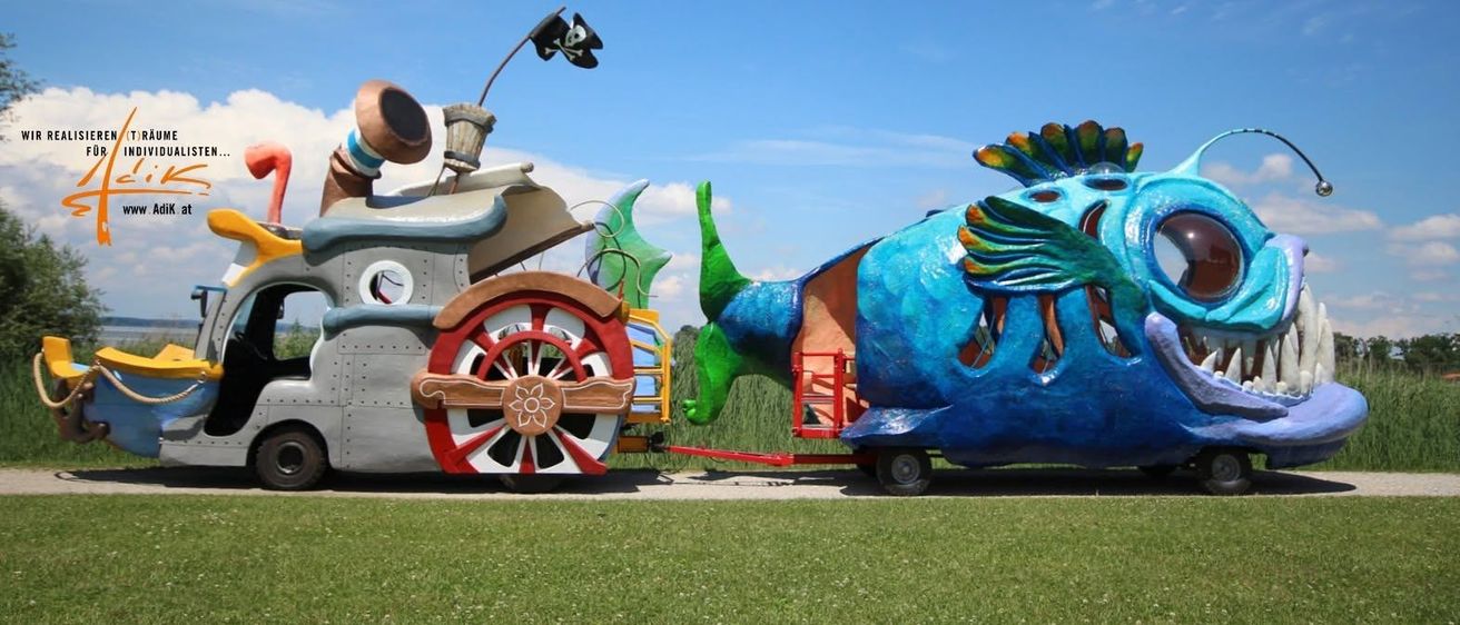 Ein farbenfrohes Piratenschiff und ein fischförmiges Fahrzeug mit einer roten Reling und einer schwarzen Piratenflagge sind auf der Straße geparkt. Der Himmel ist blau mit einigen Wolken.