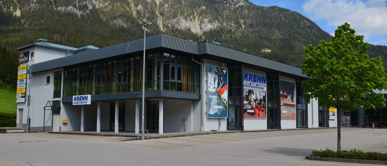 Die Vorderseite eines modernen Gebäudes namens Krenn, gelegen in einer Bergregion. Das Gebäude verfügt über große Glasfenster und Banner, die für Krenn-Sport werben.