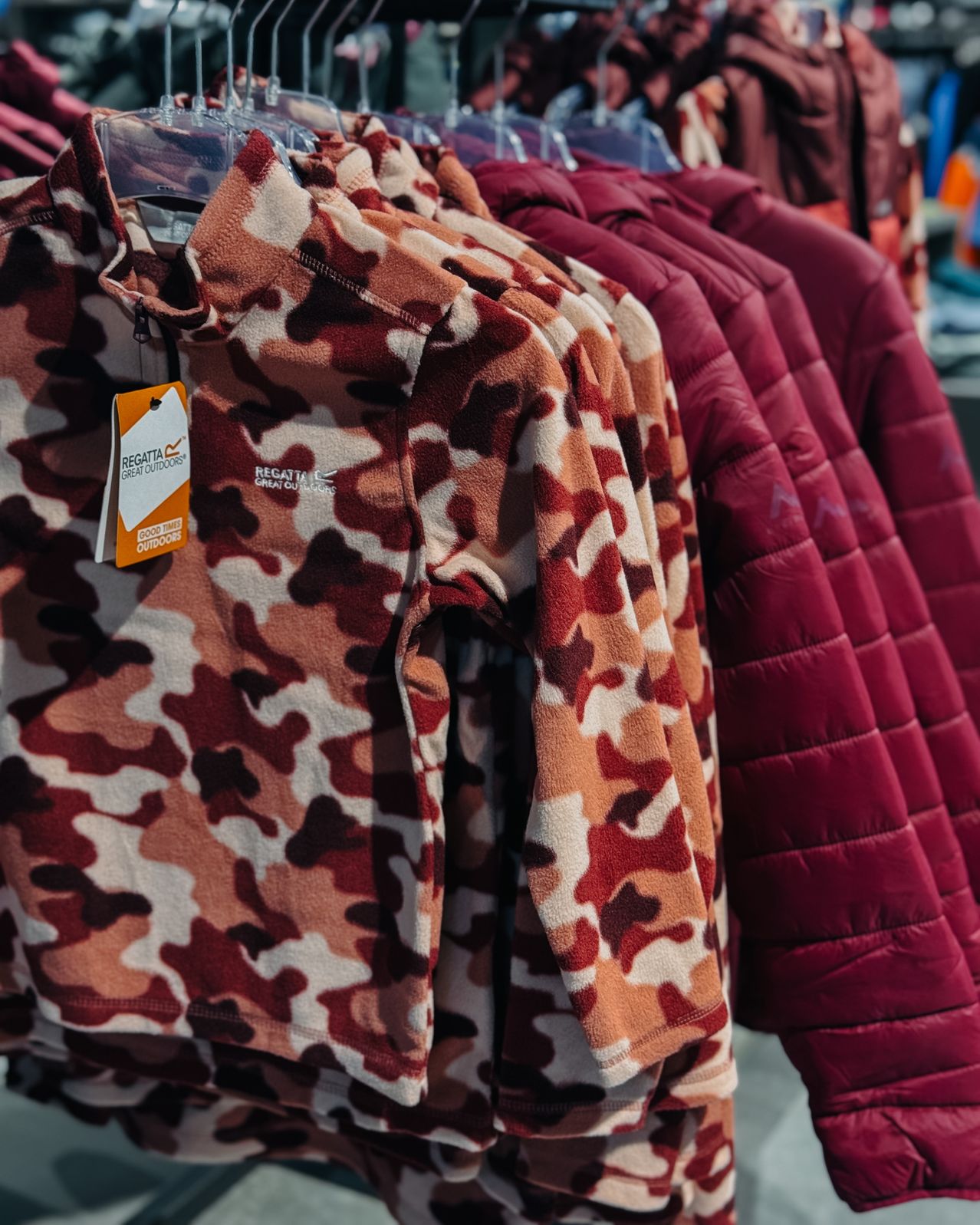 Eine Reihe von Jacken an einem Kleiderständer, darunter eine Tarnjacke von Regatta Great Outdoors mit Etikett, neben einer schlichten Weinroten Jacke.
