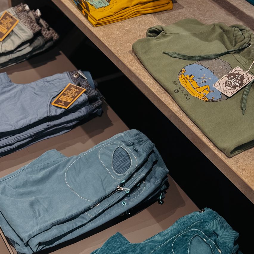Eine Kleidungsdisplay mit ordentlich gefalteten Jeans und einer Kapuzenjacke auf einer grauen Oberfläche. Die Jeans haben einzigartige Designelemente und Etiketten, während die Kapuzenjacke ein Cartoonbild und ein Etikett hat.