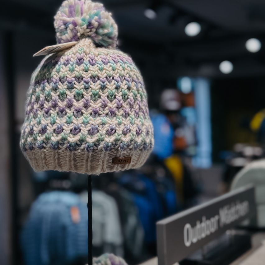 Eine bunte gestrickte Mütze mit Bommel wird in einem Geschäft ausgestellt, mit verschwommener Kleidung und einem Schild mit der Aufschrift Outdoor Watch im Hintergrund.