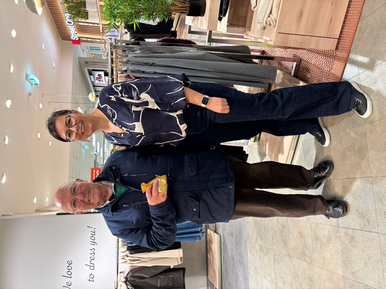 A smiling couple stands inside a clothing store. The woman wears glasses and a blue blouse. The man holds a yellow object. They are surrounded by hanging clothes.