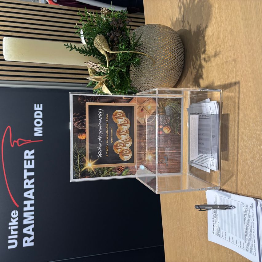 A clear box with a Christmas card displaying gold coins sits on a wooden surface. A plant in a decorative vase and a candle are behind it. A red arrow sign with the text 'Ulrike RAMHARTER MODE' is on the left. Papers are on the right.