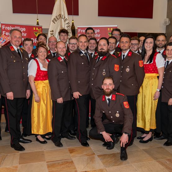 Eine Gruppe Feuerwehrleute in Uniform, einige in traditioneller Tracht, posieren für ein Foto mit Bannern im Hintergrund.