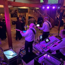 Eine Band spielt in einer Location mit lila Beleuchtung. Musiker spielen auf der Bühne mit Instrumenten und Mikrofon. Das Publikum tanzt auf dem Boden.