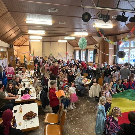 Eine festliche Indoor-Versammlung mit Tischen, Stühlen, Ballons und bunten Dekorationen. Viele Menschen, einschließlich Kinder in Kostümen, sitzen oder stehen. Flaschen, Teller und andere Gegenstände sind auf den Tischen.