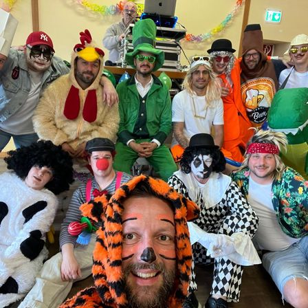 Eine Gruppe von Erwachsenen in verschiedenen Kostümen, darunter ein Huhn, ein grüner Elf, ein Clown und ein Tiger, posiert für ein Foto in einem festlich dekorierten Raum.
