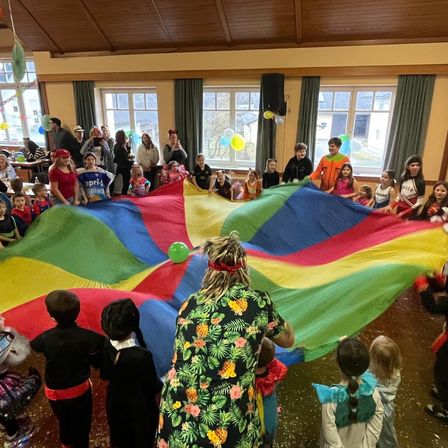Eine Gruppe von Kindern und Erwachsenen versammelt sich um einen großen mehrfarbigen Fallschirm in einem Saal mit großen Fenstern und Dekorationen. Einige tragen Kostüme.