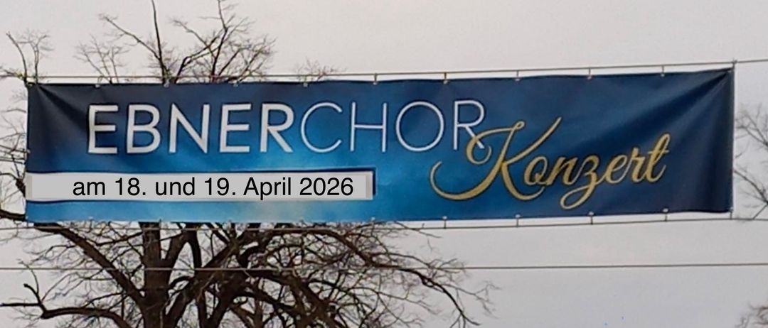 Ein blauer Banner zeigt den Text 'WebnerChor Konzert' mit den Daten 18. und 19. April 2026. Der Banner ist an einem Metallrahmen über einem kahlen Baum befestigt.
