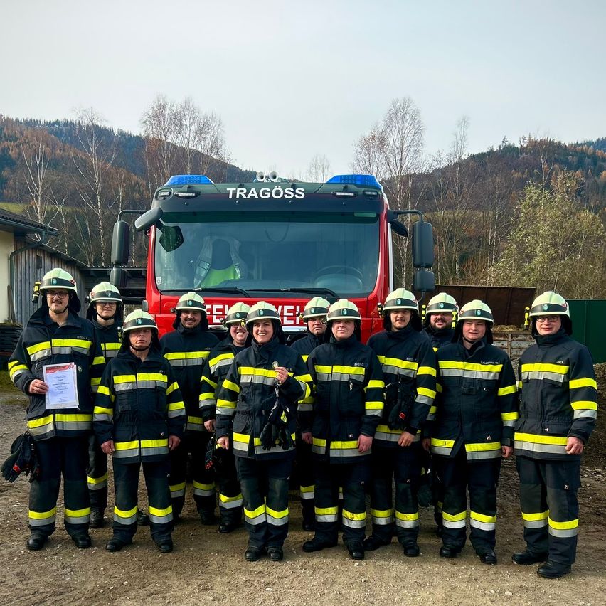 A group of firefighters in uniform poses in front of a red Tragöss fire truck with a mountain range in the background.
