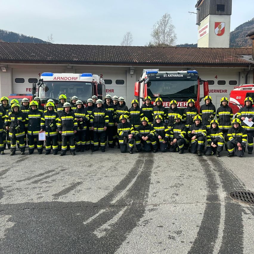 A group of firefighters in yellow and black uniforms poses in front of two fire trucks outside a fire station.