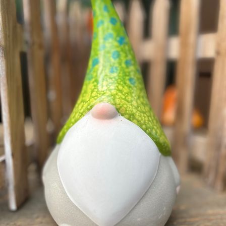 Eine kleine, handbemalte Gnomfigur mit einem weißen Körper und einem grünen Hut mit blauen Punkten. Der Gnom steht vor einem Holzzaun.
