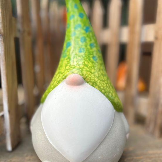 Eine kleine, handbemalte Gnomfigur mit einem weißen Körper und einem grünen Hut mit blauen Punkten. Der Gnom steht vor einem Holzzaun.