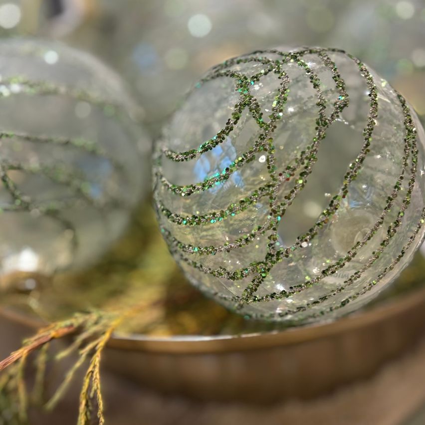 Eine Nahaufnahme von zwei Glas-Weihnachtskugeln mit grünen und silbernen Verzierungen auf einer metallischen Oberfläche. Der Hintergrund ist leicht verschwommen.