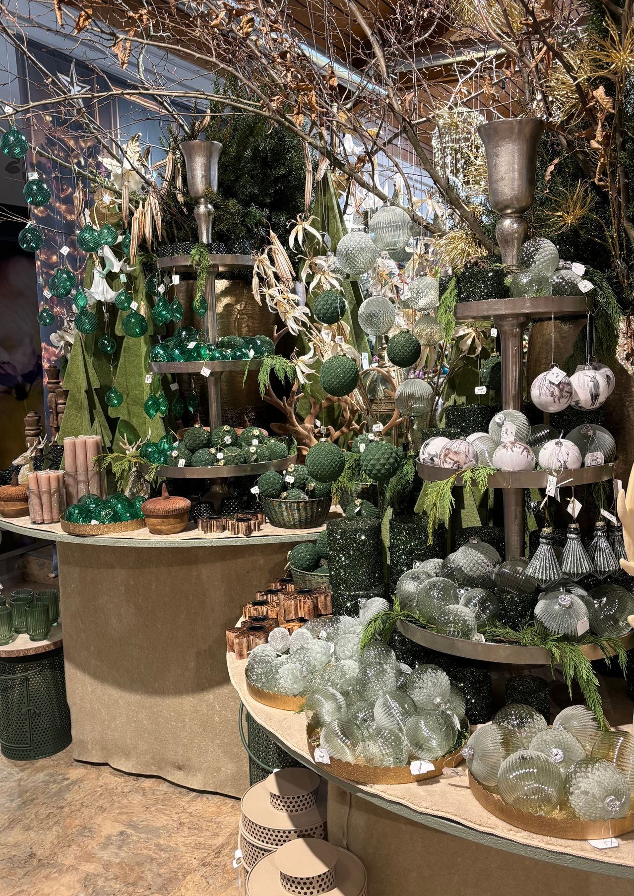 Eine Ausstellung von grünen und silbernen Glaskugeln, dekorativen Kerzen und Grünpflanzen. Eine Vielzahl von Dekorationen auf einem dreistufigen Ständer, während ein Tisch Kerzen und Eicheln hält.