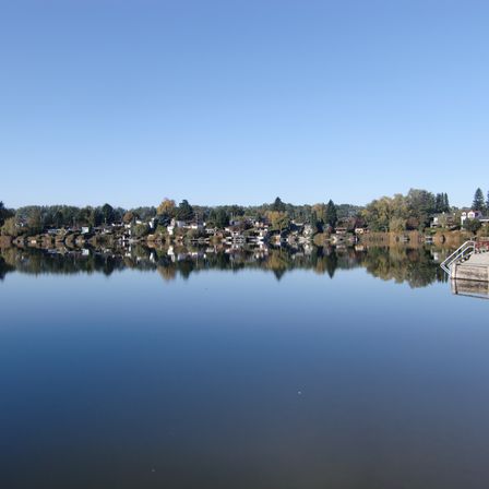 Ein ruhiger See, der die Stadt mit Häusern und Bäumen am Ufer unter einem klaren blauen Himmel widerspiegelt.
