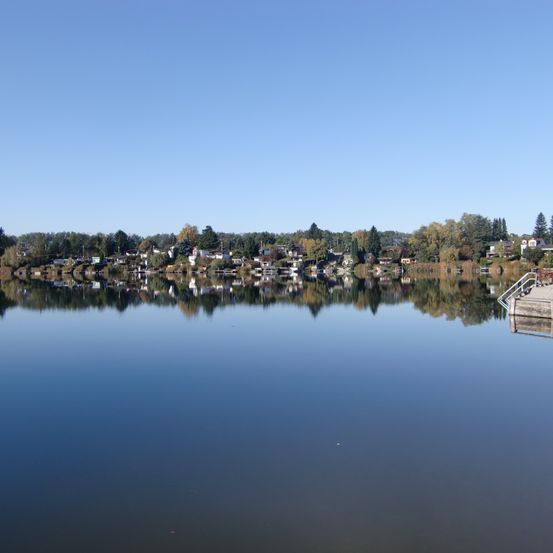 Ein ruhiger See, der die Stadt mit Häusern und Bäumen am Ufer unter einem klaren blauen Himmel widerspiegelt.