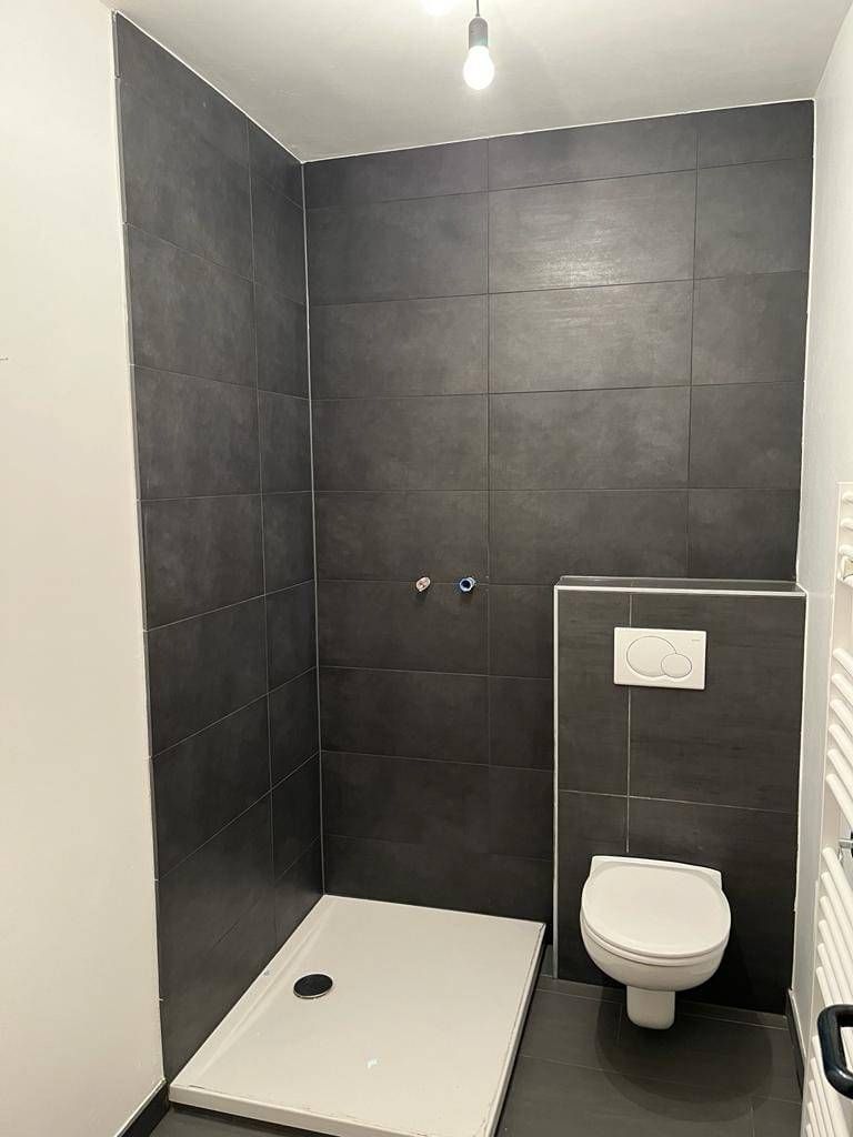 Ein modernes Badezimmer mit einer grauen gefliesten Duschkabine und Boden, einem weißen WC und einem weißen Heizkörper an der Wand.