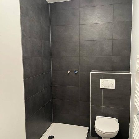 Ein modernes Badezimmer mit einer grauen gefliesten Duschkabine und Boden, einem weißen WC und einem weißen Heizkörper an der Wand.