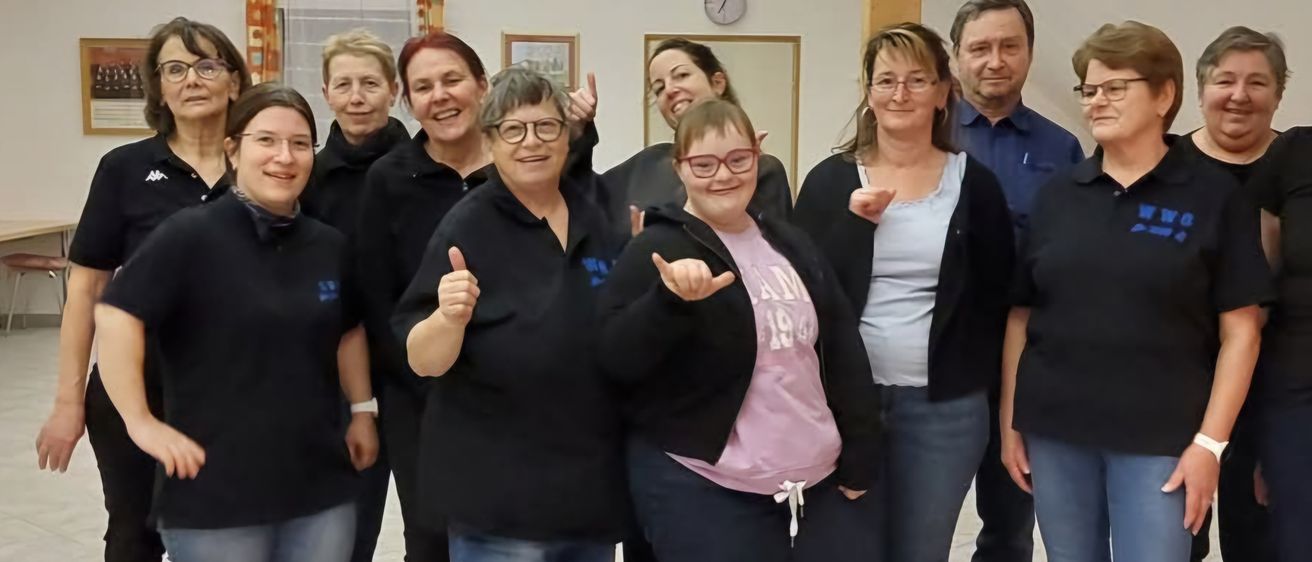 Eine Gruppe Erwachsener, darunter eine Frau mit Brille und einem rosa Shirt, posiert für ein Foto in einem Raum.